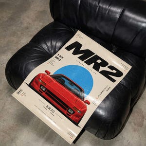 Könnte beinhalten: Ein rechteckiges Poster mit einem roten Toyota MR2 Sportwagen, der vor einem blauen Kreis zentriert ist. Der Text lautet MR2, Legend Lives On, SW20, 1990-1999 und enthält japanische Schriftzeichen. Das Poster hat einen cremefarbenen Hintergrund.