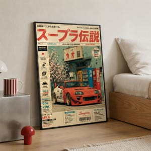 Könnte beinhalten: Ein gerahmtes Poster mit einem roten Toyota Supra MK4 Sportwagen, der vor einem Gebäude mit Kirschblüten geparkt ist. Das Design enthält japanischen Text, technische Spezifikationen, einen Strichcode und den Titel Supra Legends im Stil eines Magazins.
