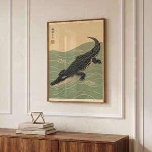 May include: A framed wall art print featuring a dark black crocodile swimming through stylised green water waves. The background is a light beige colour with Japanese text in the upper left corner and a small red square seal stamp below it.