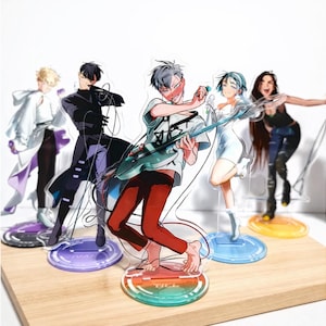 May include: Acrylic anime character figures of a band, each on a circular base. The figures are of a man playing a guitar, a woman with a microphone, and other characters in various poses. The figures are on a wooden surface.