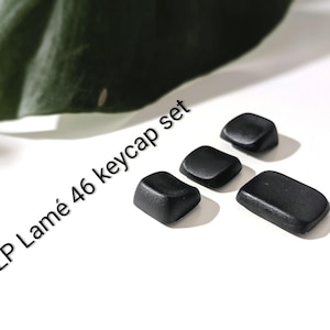 Op de afbeelding: Een set van vier zwarte keycaps in verschillende vormen, waaronder vierkant en rechthoekig, wordt weergegeven op een wit oppervlak. De tekst "KLP Lamé 46 keycap set" is diagonaal in het zwart gedrukt.