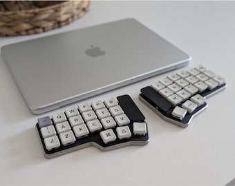 May include: A silver laptop with an Apple logo sits next to a split ergonomic mechanical keyboard. The keyboard consists of two separate black and silver units featuring white keycaps with black letters, numbers, and symbols arranged in a non-standard layout.