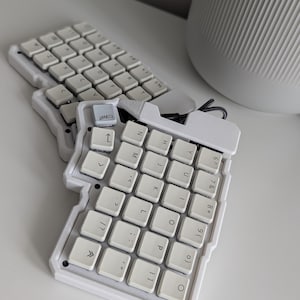 Może przedstawiać: Dzielona ergonomiczna klawiatura mechaniczna z dwiema białymi, wyprofilowanymi obudowami i białymi klawiszami. Klawisze mają czarne litery, cyfry i symbole. Jeden klawisz w lewym górnym rogu prawej części posiada małą szarą ikonę.