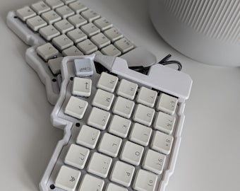 May include: A split ergonomic mechanical keyboard featuring two white, contoured cases with white keycaps. The keys display standard letters, numbers, and symbols in black. One key in the top left corner of the right half features a small gray icon.
