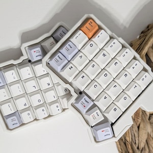 Puede incluir: Un teclado mecánico ergonómico dividido con teclas blancas y un chasis blanco. Las teclas presentan letras negras, y algunas muestran texto como After, Hyper, Toggle y Final. Una tecla es de color naranja con el texto NS 03-01.