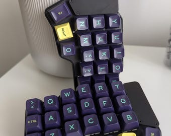 May include: A split ergonomic mechanical keyboard half featuring a black chassis and dark purple keycaps with light green lettering. The layout includes specific keys labeled RIGHT, DOWN, CODE, CTRL, SHIFT, and FN, with a unique staggered key arrangement.