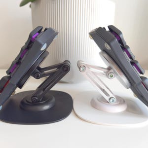 Op de afbeelding: Twee mechanische toetsenbordhelften gemonteerd op verstelbare standaards. De ene standaard is matzwart en de andere is zilverkleurig. Elke toetsenbordhelft heeft donkergrijze toetsen met paarse accenten die tussen de toetsen zichtbaar zijn. De standaards hebben scharnierende armen.