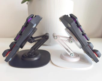 May include: Two mechanical keyboard halves mounted on adjustable stands. One stand is matte black and the other is silver. Each keyboard half features dark gray keycaps with purple accents visible between the keys. The stands have articulated arms.