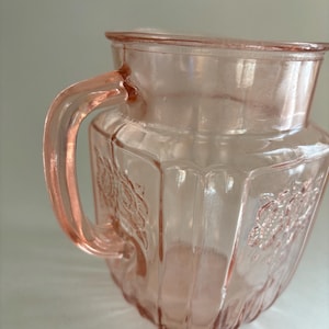 May include: A vintage pink glass pitcher with a handle. The pitcher has a faceted design and floral detailing. The pitcher is made of transparent glass and is likely used for serving beverages. The pitcher is a light pink color.