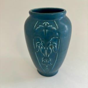 May include: A teal-colored ceramic vase with a detailed embossed floral design. The vase has a rounded body and a slightly flared rim. The embossed design is centered on the front of the vase.