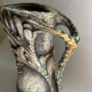 May include: A detailed close-up of a ceramic pitcher with a textured surface. The pitcher features a handle and is decorated with a speckled pattern in shades of gray, black, and cream, with accents of gold and green.