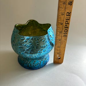 May include: A small, iridescent blue and green glass bowl with a textured, bubbled surface and a scalloped rim. A wooden ruler is positioned vertically next to the bowl, showing a height of approximately 3.5 inches for the glass object.