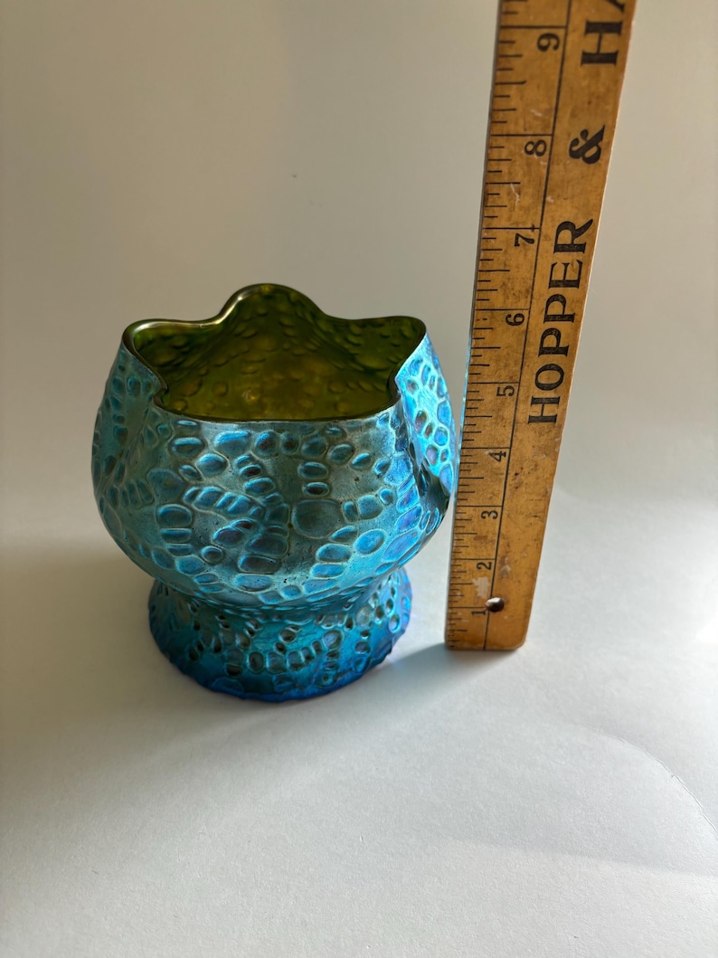 May include: A small, iridescent blue and green glass bowl with a textured, bubbled surface and a scalloped rim. A wooden ruler is positioned vertically next to the bowl, showing a height of approximately 3.5 inches for the glass object.