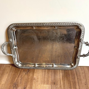 May include: A rectangular silver-toned serving tray with a polished reflective surface, featuring an ornate embossed border pattern and two decorative curved handles on the sides. The center of the tray displays a faint, etched floral design.