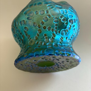 May include: A glass vase with an iridescent finish in shades of teal, blue, and green. The surface features a raised, textured pattern of interconnected circular and organic shapes that create a tactile, honeycomb-like appearance across the body.