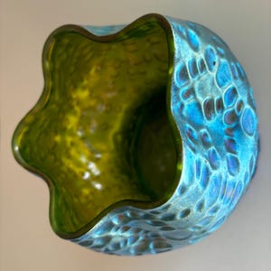 May include: A glass bowl with a wavy, scalloped rim. The exterior features a textured, dimpled surface with an iridescent blue finish, while the interior is a smooth, translucent lime green color. The bowl has a rounded, bulbous shape.