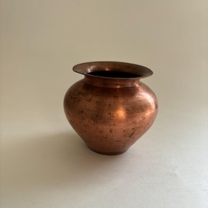 May include: A small, rounded copper vessel with a wide, flared rim and a narrow base. The metal surface displays a warm, reddish-brown metallic tone with visible natural oxidation, patina, and subtle horizontal texture lines around the body.
