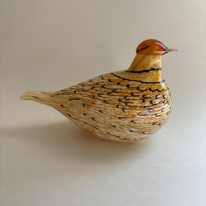 May include: A glass bird figurine featuring a rounded body with a speckled pattern of amber, white, and black. The head is a solid translucent amber color with a pointed beak, and the tail tapers to a point, showcasing a smooth, polished finish.