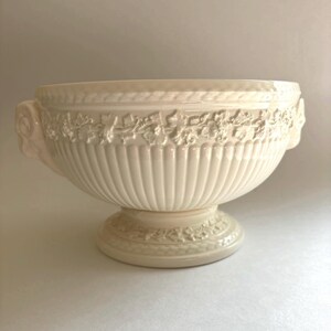 May include: A cream-colored ceramic bowl with a pedestal base and two side handles. The exterior features a raised relief pattern of grapevines and leaves around the rim and base, with vertical fluting along the body of the bowl.