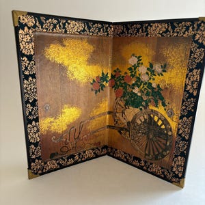 May include: A two-panel folding screen featuring a gold leaf background with a wooden cart carrying red and white flowers. The border is black fabric with a gold floral pattern, and the corners are reinforced with small, metallic gold-toned accents.