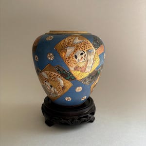 May include: A blue ceramic vase featuring gold-toned fan-shaped panels with illustrations of figures, accented by white dot patterns. The vase rests on a dark brown, carved wooden stand with a circular base and decorative feet.
