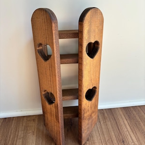 May include: A vertical wooden rack with a dark brown finish, featuring two tall side panels connected by three horizontal shelves. Each side panel has two cut-out heart shapes, creating a symmetrical design with open spaces between the shelves.