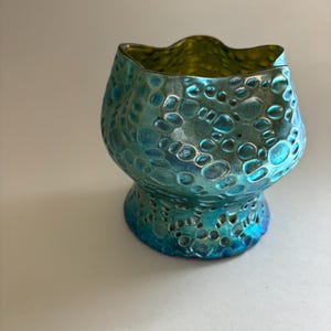May include: A small glass vessel with a scalloped rim and a bulbous body. The exterior features a textured, raised circular pattern with an iridescent finish that shifts between shades of metallic blue, teal, and green across the surface.