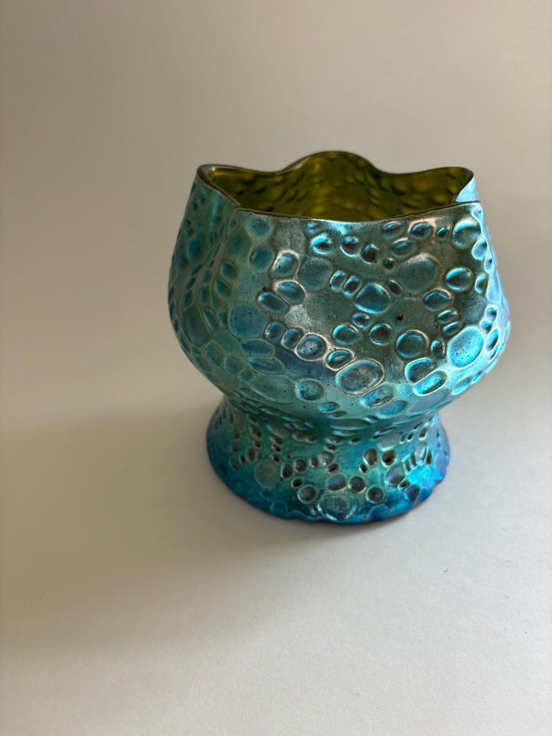 May include: A small glass vessel with a scalloped rim and a bulbous body. The exterior features a textured, raised circular pattern with an iridescent finish that shifts between shades of metallic blue, teal, and green across the surface.