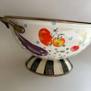 May include: A white enameled metal colander featuring a hand-painted vegetable motif including a purple eggplant, red tomatoes, and a radish. The bowl has a brass-toned rim and handles, with a base decorated in black and white vertical stripes.