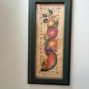 May include: A rectangular wall art piece in a dark green wooden frame featuring a vertical illustration of stacked fruit including a pear, apple, peach, grapes, and a watermelon slice, accented with yellow sunflowers on a textured tan background.