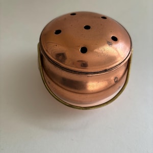 May include: A copper-colored, round container with a lid featuring several small holes. The container has a brass handle and a slightly aged appearance, suggesting it is vintage or antique. The design is simple and functional.