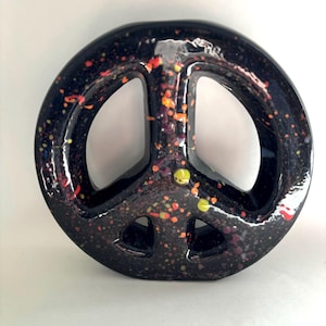 May include: A circular ceramic peace sign sculpture with a glossy black finish. The surface is decorated with a speckled pattern of small, vibrant dots in shades of orange, yellow, and red, creating a textured appearance across the entire object.
