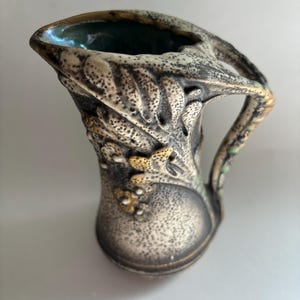 May include: A small, antique-style pitcher with a textured, speckled surface. The pitcher features a handle and is decorated with embossed leaf and floral patterns in shades of beige, brown, and gold. The interior is a deep teal color.
