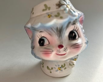 Puede incluir: Una figura de gato de cerámica con base blanca y detalles florales en azul y verde. El gato tiene grandes ojos negros, mejillas rosadas y una pequeña nariz roja. Lleva un sombrero blanco a juego con flores azules y hojas verdes pintadas.