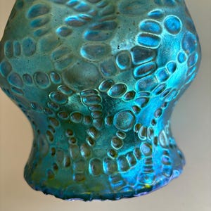 May include: A turquoise glass vase featuring an iridescent finish and a raised, textured pattern of interconnected circular and organic shapes. The surface displays a metallic sheen with shifting blue and green tones across the entire exterior.