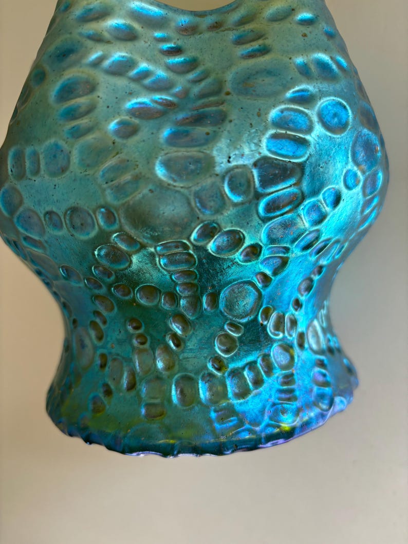 May include: A turquoise glass vase featuring an iridescent finish and a raised, textured pattern of interconnected circular and organic shapes. The surface displays a metallic sheen with shifting blue and green tones across the entire exterior.
