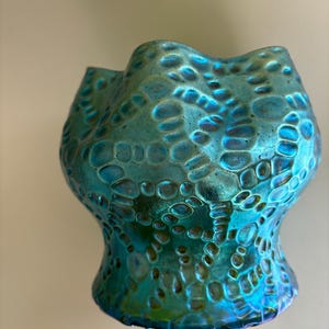 May include: A small, iridescent glass vase featuring a vibrant teal and blue color palette. The surface is covered in a raised, textured pattern of interconnected circular and oval shapes, creating a honeycomb-like effect with a wavy, scalloped rim.