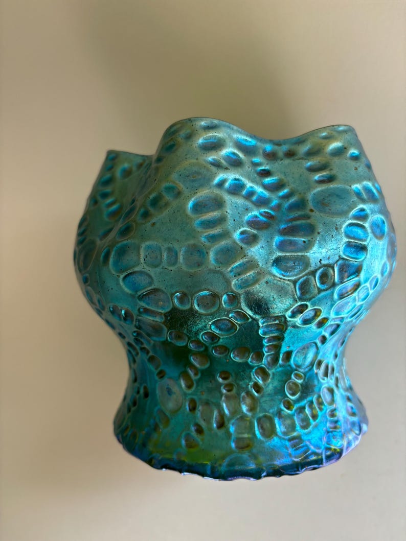 May include: A small, iridescent glass vase featuring a vibrant teal and blue color palette. The surface is covered in a raised, textured pattern of interconnected circular and oval shapes, creating a honeycomb-like effect with a wavy, scalloped rim.