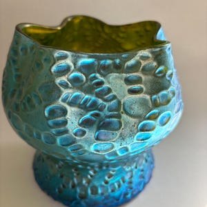 May include: A small, iridescent glass bowl featuring a textured, pebble-like surface pattern. The exterior displays a vibrant blue and teal finish with a metallic sheen, while the interior reveals a contrasting lime green hue and a wavy rim.