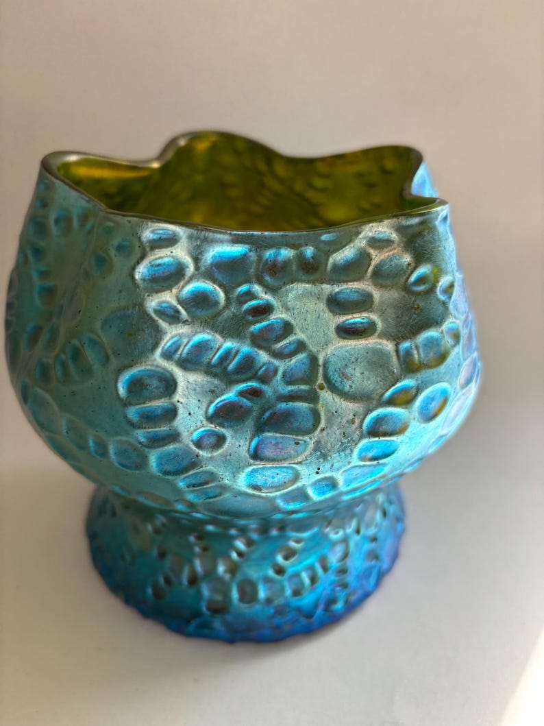 May include: A small, iridescent glass bowl featuring a textured, pebble-like surface pattern. The exterior displays a vibrant blue and teal finish with a metallic sheen, while the interior reveals a contrasting lime green hue and a wavy rim.