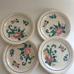 May include: Four off-white ceramic plates with a floral design. Each plate features a pink rose, blue and yellow flowers, and green leaves. A blue line borders the edge of each plate.