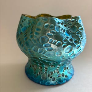 May include: A teal and blue glass vessel with a scalloped rim and a textured surface featuring raised, rounded, bubble-like patterns. The exterior has a metallic, iridescent finish that shifts in color, while the interior is smooth and gold.