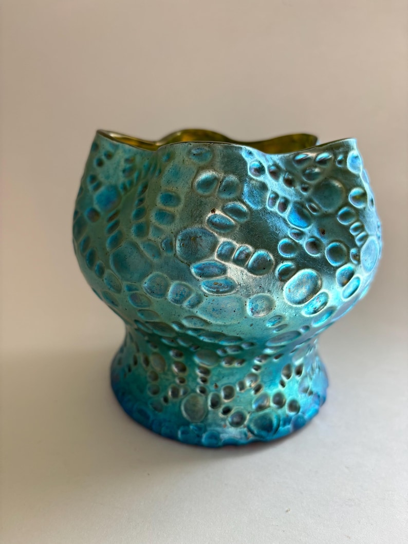 May include: A teal and blue glass vessel with a scalloped rim and a textured surface featuring raised, rounded, bubble-like patterns. The exterior has a metallic, iridescent finish that shifts in color, while the interior is smooth and gold.