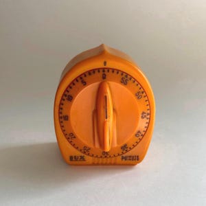 May include: A bright orange Lux Minute Minder kitchen timer with a rounded, arch-like shape. The face features a central rotating dial with a vertical indicator, surrounded by black printed numbers from 0 to 55, marking minutes in five-minute increments.