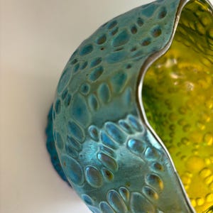 May include: A glass bowl featuring a textured exterior with a metallic turquoise finish and a smooth, vibrant lime green interior. The rim has an irregular, wavy shape, and the outer surface is covered in raised, circular, bubble-like indentations.