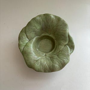May include: A ceramic bowl shaped like a green cabbage head with detailed, textured leaves radiating from a central circular depression. The piece features varying shades of sage and olive green with a matte finish, mimicking the appearance of a vegetable.