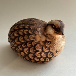May include: A small, rounded bird figurine crafted with a light tan base color and dark brown, scalloped feather patterns across its body. The bird has a smooth, rounded head with a dark brown eye marking and a small, pointed beak facing forward.