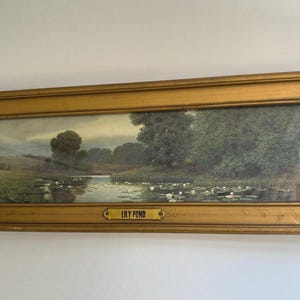 May include: A rectangular landscape painting titled LILY POND, featuring a calm body of water with white lily pads, surrounded by green trees and grassy banks under a cloudy sky, enclosed in a wide, distressed gold-toned wooden frame.
