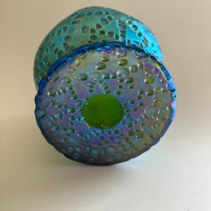 May include: A round glass object with an iridescent blue and green finish featuring a textured surface of raised circular bumps. The base is flat and circular with a smooth, solid green center surrounded by the same textured, iridescent pattern.