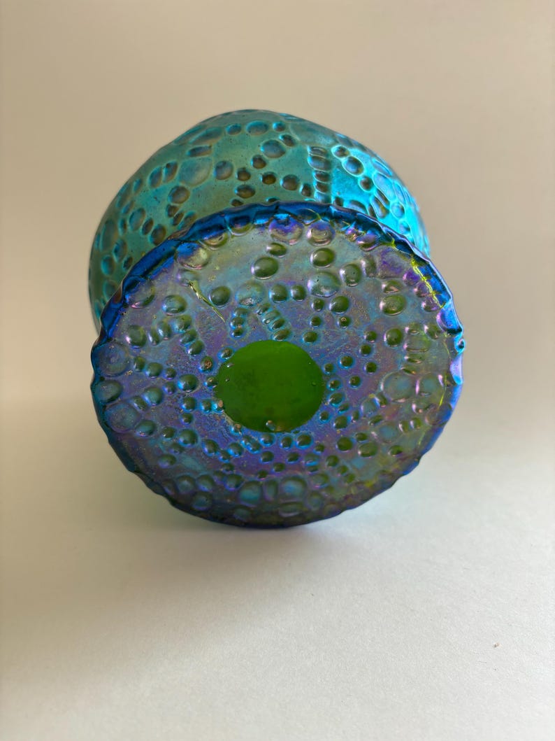 May include: A round glass object with an iridescent blue and green finish featuring a textured surface of raised circular bumps. The base is flat and circular with a smooth, solid green center surrounded by the same textured, iridescent pattern.